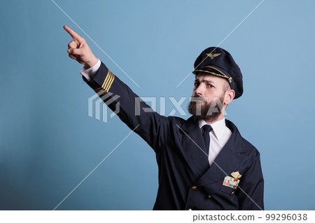 Serious captain pointing at sky with index finger, wearing professional aviation uniform, plane pilot looking at camera. Confident aviation academy aviator standing, studio medium shot Serious captain pointing at sky with index finger, wearing professional aviation uniform, plane pilot looking at camera. Confident aviation academy aviator standing, studio medium shot 99296038
