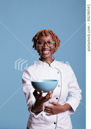 African american woman happily smiling, professional chef uniformed holding blue bowl, vertical studio portrait. Happy cooker showing a container, looking at the camera. African american woman happily smiling, professional chef uniformed holding blue bowl, vertical studio portrait. Happy cooker showing a container, looking at the camera. 99296041