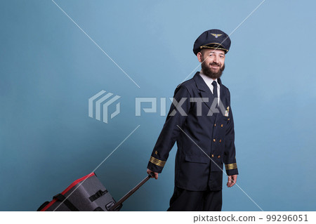 Airplane pilot in professional airline uniform standing with baggage, looking at camera. Ready pilot with luggage waiting for flight in airport, aviator holding luggage 99296051