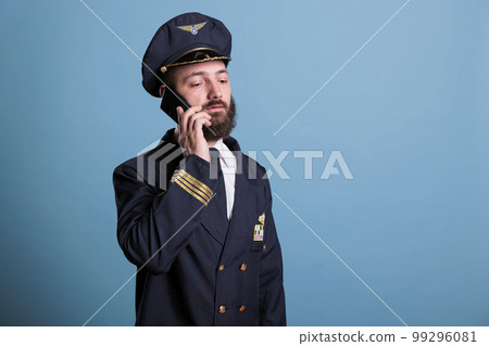 Middle aged pilot talking on smartphone, holding mobile phone, having conversation. Airlane captain in uniform with calm neutral facial expression answering telephone call Middle aged pilot talking on smartphone, holding mobile phone, having conversation. Airlane captain in uniform with calm neutral facial expression answering telephone call 99296081