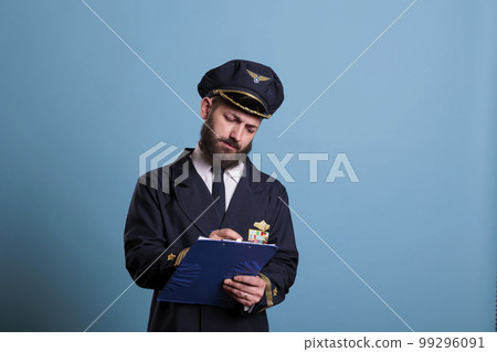 Airplane captain holding clipboard, concentrated pilot writing papers. Aircraft aviator in professional aviation uniform filling form in airport front view, studio medium shot on blue background Airplane captain holding clipboard, concentrated pilot writing papers. Aircraft aviator in professional aviation uniform filling form in airport front view, studio medium shot on blue background 99296091