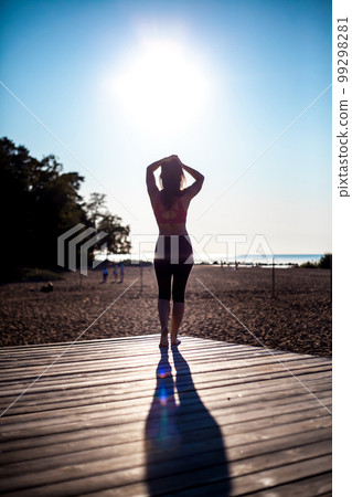 backwards view as a slim girl after fitness exercises walks on a wooden platform on a sunny summer sea beach 99298281