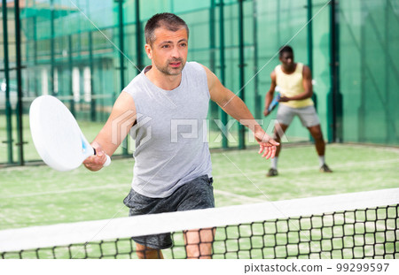 Male players playing padel in a padel court outdoor behind net Male players playing padel in a padel court outdoor behind net 99299597