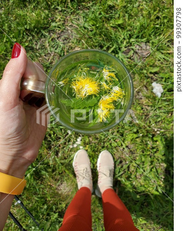 A woman holding a glass mug of herbal tea with dandelion on the grass on a sunny day 99307798