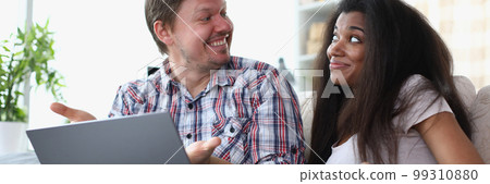 Interethnic family couple laughing and chatting while sitting on sofa at home with laptop. 99310880