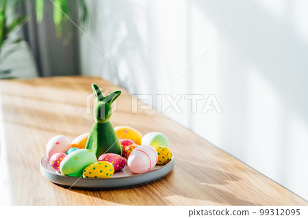 Easter home decor. Green bunny rabbit figurine and colored easter chocolate eggs on plate on wooden console with green home plants and sunlight and shadows on white wall. Selective focus. Copy space. Easter home decor. Green bunny rabbit figurine and colored easter chocolate eggs on plate on wooden console with green home plants and sunlight and shadows on white wall. Selective focus. Copy space. 99312095