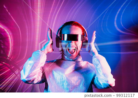 Portrait of emotionally shouting woman in futuristic sunglasses and headphones in pink and blue neon light. Music lover. Silent disco. Woman on background of music vibes background. Futuristic Party 99312117