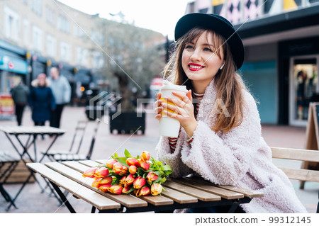 Stylish young smiling woman enjoying coffee from a reusable cup, listening to music, podcast using wireless earphones in a street cafe. Fresh tulips bouquet on the table. Springtime street fashion Stylish young smiling woman enjoying coffee from a reusable cup, listening to music, podcast using wireless earphones in a street cafe. Fresh tulips bouquet on the table. Springtime street fashion 99312145