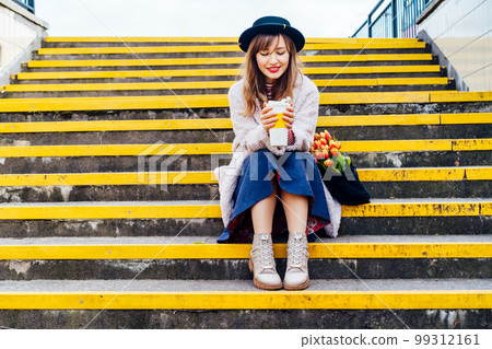 Portrait of young smiling fashion woman with fresh tulips in her net bag holding reusable coffee cup while sitting on stairs at the city street. Urban street fashion. Springtime mood. Selective focus Portrait of young smiling fashion woman with fresh tulips in her net bag holding reusable coffee cup while sitting on stairs at the city street. Urban street fashion. Springtime mood. Selective focus 99312161