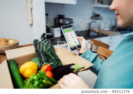 Online home food delivery. Focus on active app with order list on the phone screen. Woman checking her order. Cardboard box with fresh vegetables standing on the kitchen table. Local farmer food. 99312176