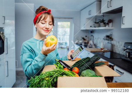 Online home food delivery. Woman checking her online order list on her phone. Cardboard box with fresh vegetables and fruits standing on the kitchen table. Local farmer food. Start of a healthy life 99312180