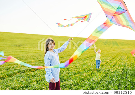 Smiling girl and brother boy running with flying colorful kites on the high grass meadow. Happy childhood moments 99312510