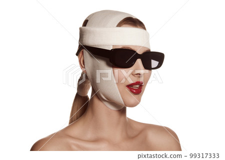 Female face bandaged with medical bandages and sunglasses with human emotions. Making beauty, modifying face to make surgical correction, plastic surgery. correction of asymmetry. 99317333