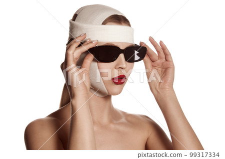 Female face bandaged with medical bandages and sunglasses with human emotions. Making beauty, modifying face to make surgical correction, plastic surgery. correction of asymmetry. 99317334