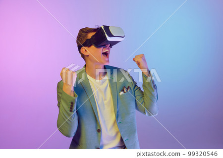 Futurism, virtual reality. Young handsome man in stylish jacket posing in VR glasses over purple background in neon light. Concept of emotions, fashion and lifestyle 99320546