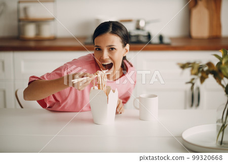 Portrait of happy smiling girl sitting in the kitchen. Cute young woman in casual clothes eats noodles from paper box with chopsticks. Delivery of ready-made fast food at home. Asian cuisine. Portrait of happy smiling girl sitting in the kitchen. Cute young woman in casual clothes eats noodles from paper box with chopsticks. Delivery of ready-made fast food at home. Asian cuisine. 99320668