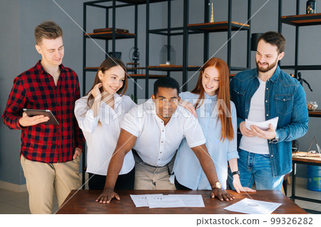 Confident African-American business team leader stading surrounded by colleagues and looking confidently at camera. Young creative business team posing near desk in modern office room. Confident African-American business team leader stading surrounded by colleagues and looking confidently at camera. Young creative business team posing near desk in modern office room. 99326282