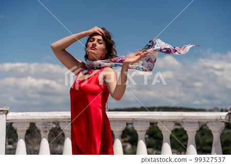 A woman in a red dress against a blue sky and white clouds with a developing scarf around her neck. She turned her face to the wind and posed for the camera. A woman in a red dress against a blue sky and white clouds with a developing scarf around her neck. She turned her face to the wind and posed for the camera. 99327537