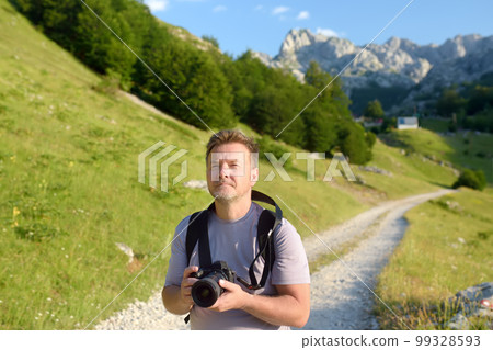 Mature man tourist with backpack and camera is hiking on mountain valley. Photographer taking a picture during travel. 99328593