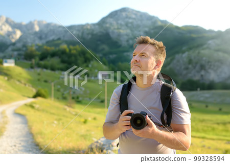 Mature man tourist with backpack and camera is hiking on mountain valley. Photographer taking a picture during travel. 99328594