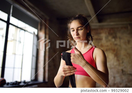 Young female boxer preparing for boxing fight.Athletic woman wearing strap on wrist before boxing practice in gym. Healthy lifestyle Young female boxer preparing for boxing fight.Athletic woman wearing strap on wrist before boxing practice in gym. Healthy lifestyle 99329002