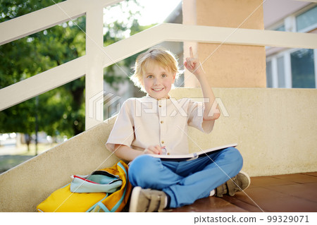 Little student doing homework on break on stair of elementary school building. Cute boy having idea and showing forefinger up. Insight concept. Back to school concept. Education for small kids. 99329071