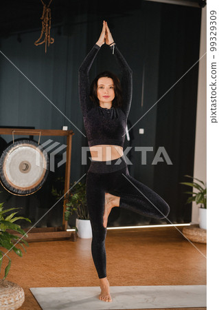 A girl in a black sports uniform does yoga standing on a mat in the gym 99329309