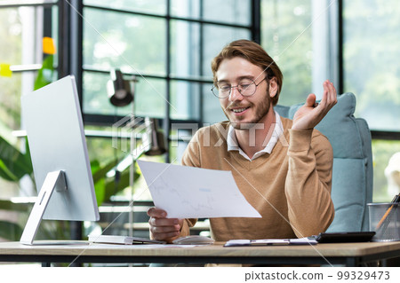 A young male student sits in the office and holds documents in his hands, studies, works at the computer, smiles. A young male student sits in the office and holds documents in his hands, studies, works at the computer, smiles. 99329473