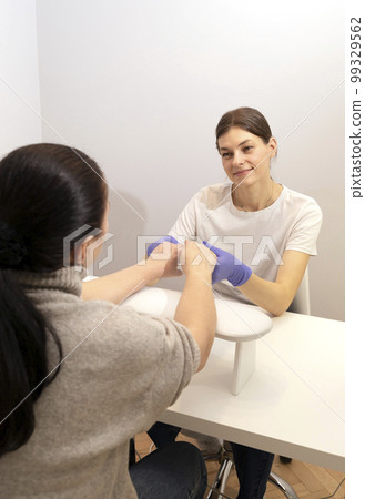 Smiling Caucasian manicure technician, nail stylist in gloves holds hands of white client, preparing Japanese eco manicure, polishing in beauty salon. Sterilized master tools in craft package on table 99329562