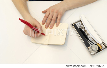 Unrecognizable nail master makes notes in notebook with pen, sitting at workstation,able in nail beauty treatment salon.Professional tools are on white table.Manicurist's routine.Top view Horizontal 99329634
