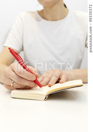 Hands of white woman make note in notebook with red pen. Cropped Caucasian female wears white shirt and marriage ring on hand, writes in a book sitting on chair at white table. Vertical plane Hands of white woman make note in notebook with red pen. Cropped Caucasian female wears white shirt and marriage ring on hand, writes in a book sitting on chair at white table. Vertical plane 99330372