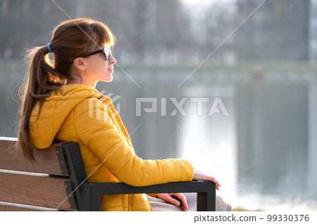 Young woman sitting on park bench relaxing on warm spring day. 99330376