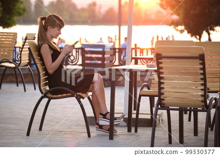 Young woman sitting at street cafe table browsing her cellphone outdoors on warm summer evening. Communication and mobile connection concept. Young woman sitting at street cafe table browsing her cellphone outdoors on warm summer evening. Communication and mobile connection concept. 99330377