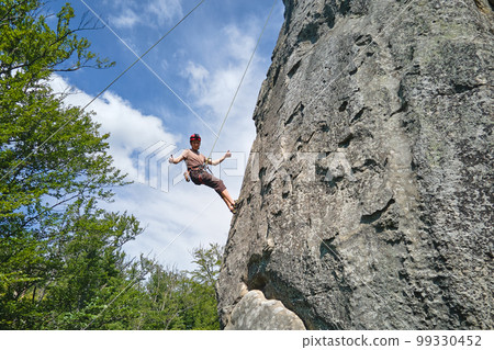 Young man climbing steep wall of rocky mountain. Male climber overcomes challenging route. Engaging in extreme sport concept. 99330452