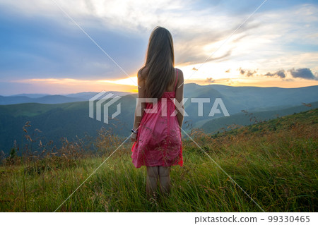 Young happy woman traveler in red dress standing on grassy hillside on a windy evening in summer mountains enjoying view of nature at sunset. Young happy woman traveler in red dress standing on grassy hillside on a windy evening in summer mountains enjoying view of nature at sunset. 99330465