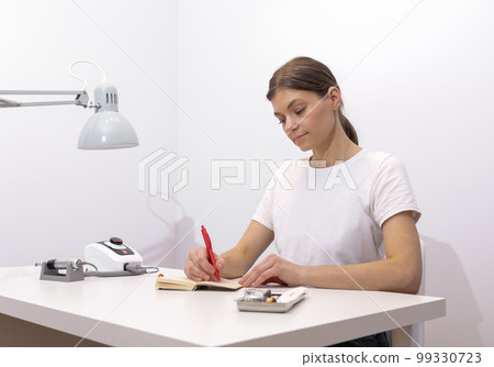 Beautiful nail beautician makes notes in notebook with pen, sitting at workstation, table in nail beauty treatment salon. Professional tools are on white table. Manicurist's routine. Horizontal plane Beautiful nail beautician makes notes in notebook with pen, sitting at workstation, table in nail beauty treatment salon. Professional tools are on white table. Manicurist's routine. Horizontal plane 99330723