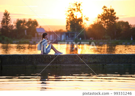 Side view of lonely woman sitting alone on lake shore on warm evening. Solitude and relaxing in nature concept. Side view of lonely woman sitting alone on lake shore on warm evening. Solitude and relaxing in nature concept. 99331246