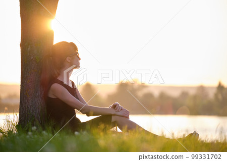 Lonely young woman sitting alone on grass lawn on lake shore enjoying warm evening. Wellbeing and relaxing in nature concept 99331702