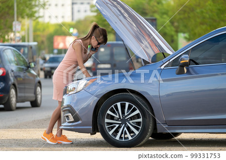 Helpless woman standing near her car with open hood takling on mobile phone calling road service for help. Young female driver having trouble with vehicle. 99331753