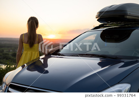 Happy young woman driver in yellow dress enjoying warm summer evening leaning on her car. 99331764
