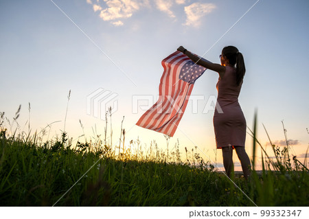 Back view of happy woman with USA national flag standing outdoors at sunset. Positive female celebrating United States independence day. International day of democracy concept. 99332347