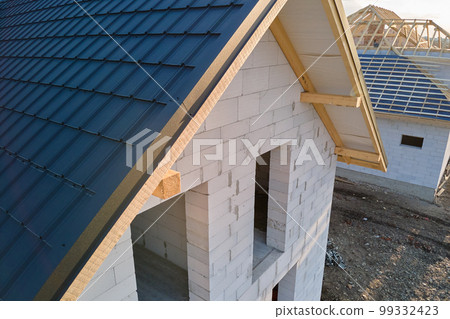 Aerial view of unfinished house with aerated lightweight concrete walls and wooden roof frame covered with metallic tiles under construction. Aerial view of unfinished house with aerated lightweight concrete walls and wooden roof frame covered with metallic tiles under construction. 99332423