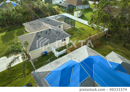 Aerial view of damaged in hurricane Ian house roof covered with blue protective tarp against rain water leaking until replacement of asphalt shingles Aerial view of damaged in hurricane Ian house roof covered with blue protective tarp against rain water leaking until replacement of asphalt shingles 99333374
