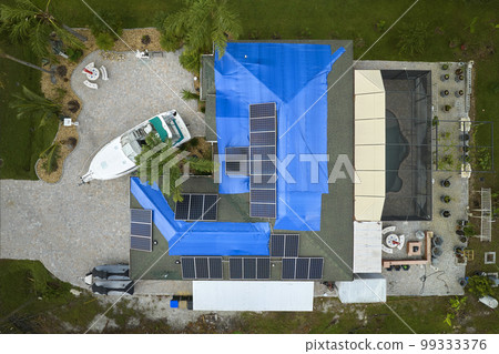 Aerial view of damaged in hurricane Ian house roof covered with blue protective tarp against rain water leaking until replacement of asphalt shingles Aerial view of damaged in hurricane Ian house roof covered with blue protective tarp against rain water leaking until replacement of asphalt shingles 99333376