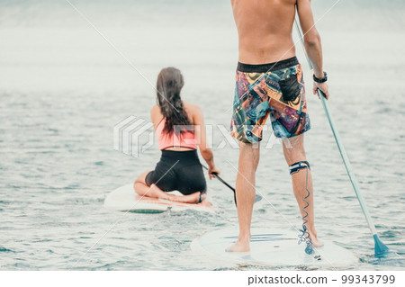 Sea woman and man on sup. Silhouette of happy young woman and man, surfing on SUP board, confident paddling through water surface. Idyllic sunset. Active lifestyle at sea or river. 99343799