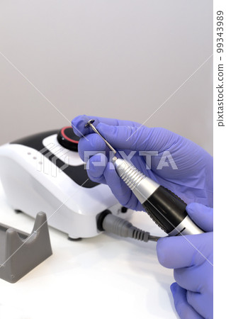 Nail beautician inserts nozzle in electric hardware drill file nail machine, wears purple groves. Nail treatment room in spa salon. Closeup, vertical. Manicure, pedicure concept. Copy space. Nail beautician inserts nozzle in electric hardware drill file nail machine, wears purple groves. Nail treatment room in spa salon. Closeup, vertical. Manicure, pedicure concept. Copy space. 99343989
