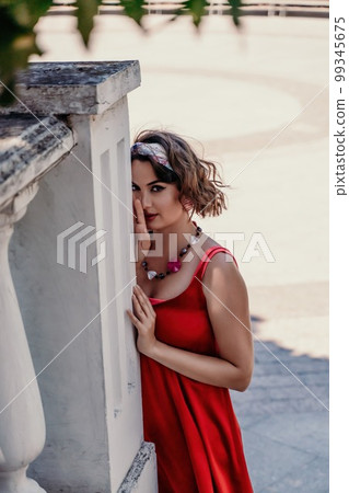 woman in a red silk dress and a bandage on her head smiles against the background of the leaves of a tree. She is leaning on the coop and looking into the camera. Vertical photo. 99345675