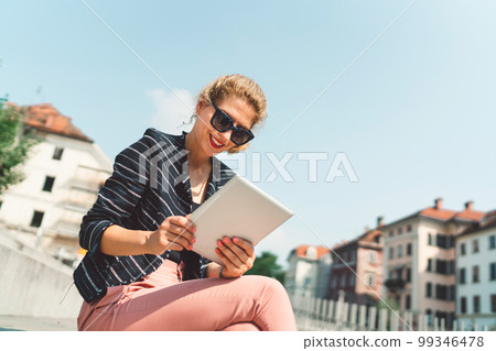 Low angle view smiling caucasian woman sitting on the sun working on her digital tablet Low angle view smiling caucasian woman sitting on the sun working on her digital tablet 99346478