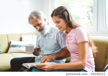 Granddaughter, young caucasian woman sitting on the couch with her grandfather reading him a book 99346521