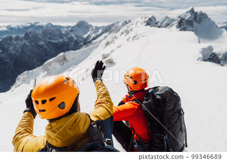 Over the head view, two mountaineers with helmets looking at the view of snowy Alps from high up Over the head view, two mountaineers with helmets looking at the view of snowy Alps from high up 99346689
